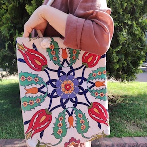Op de afbeelding: Een witte canvas tote bag met een kleurrijk bloemenontwerp met rode, groene, blauwe en gele tulpen. De tas heeft een wit touw handvat.