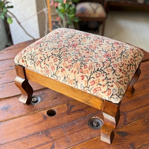 May include: A small, wooden footstool with a rectangular, cushioned top. The cushion features a floral pattern in shades of red, beige, and black. The legs are curved and the wood has a warm, brown finish.