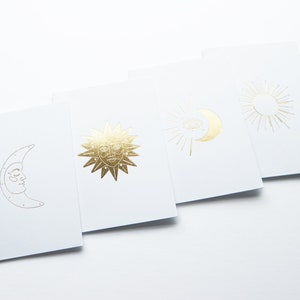 May include: Set of four white cards with gold foil designs of a crescent moon, a sun with a face, a sun with an eye, and a sun with rays.