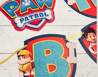 May include: A colourful banner featuring the "PAW PATROL" logo and individual letter pennants. The banner includes the letters "H" and "B" with paw print designs, and character cutouts. The banner is red, blue, yellow, and white.