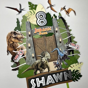 May include: A Jurassic Park-themed cake topper featuring a gate, dinosaurs, and the number 8. The topper includes the name "Shawn" in silver letters. Various dinosaur cutouts and flying pterodactyls are also included in the design.