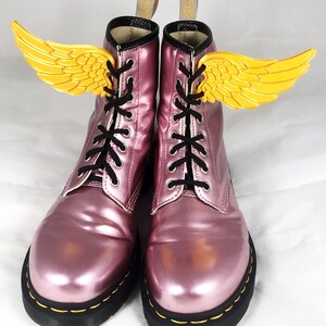 May include: A pair of pink metallic Dr. Martens boots with black laces and yellow wings attached to the laces.