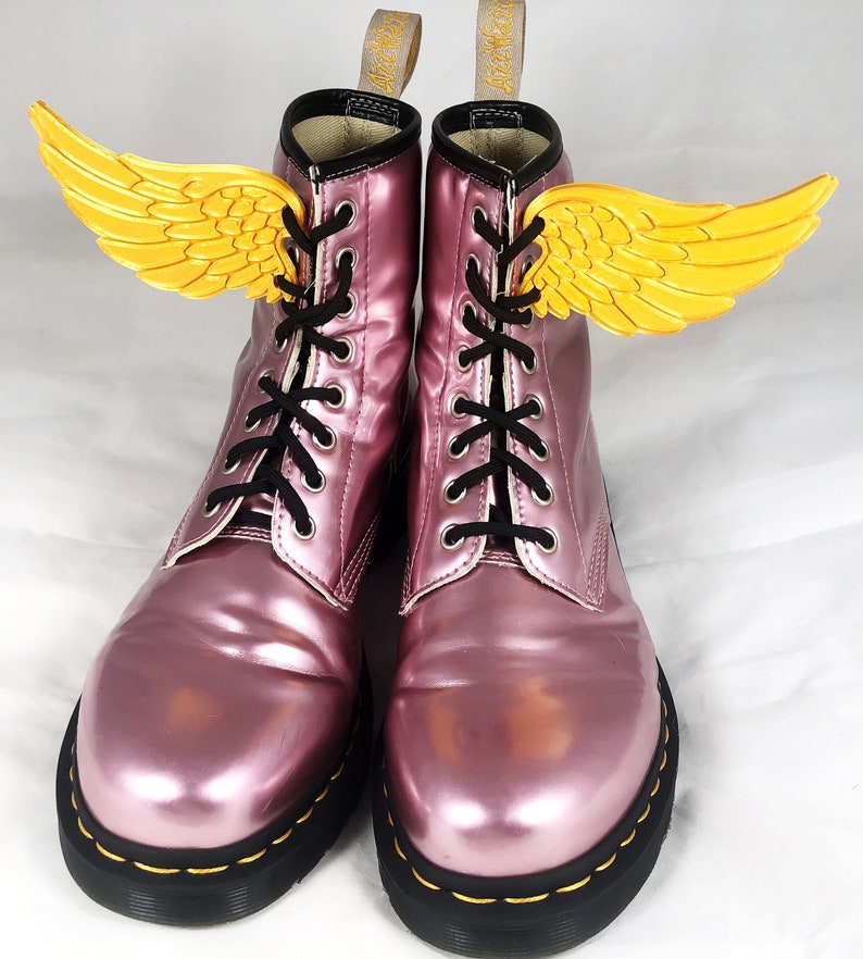 May include: A pair of pink metallic Dr. Martens boots with black laces and yellow wings attached to the laces.