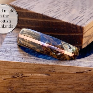 May include: A wooden ring with a copper band, in a wooden box. The ring is made of dark wood with a natural grain pattern. The box is made of light wood with a smooth finish. The text "Hand made in the Scottish Highlands" is visible in the image.