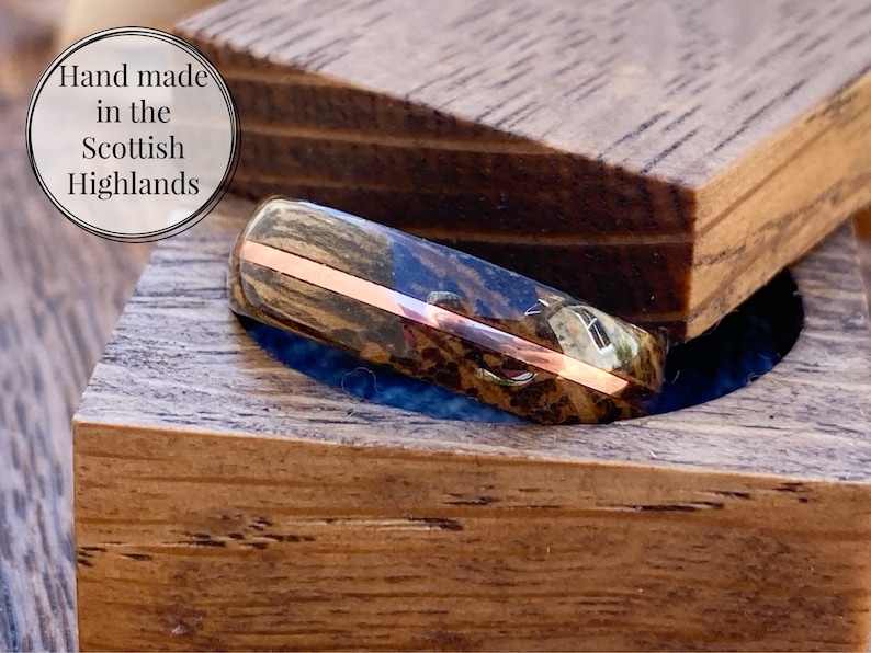 May include: A wooden ring with a copper band, in a wooden box. The ring is made of dark wood with a natural grain pattern. The box is made of light wood with a smooth finish. The text "Hand made in the Scottish Highlands" is visible in the image.