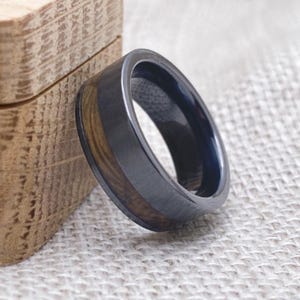 Puede incluir: Un anillo de madera negro y marr&oacute;n con una banda de metal negro. El anillo est&aacute; sobre una superficie de tela marr&oacute;n claro.