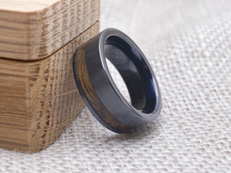 Puede incluir: Un anillo de madera negro y marr&oacute;n con una banda de metal negro. El anillo est&aacute; sobre una superficie de tela marr&oacute;n claro.