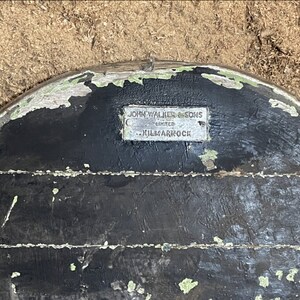 May include: A close-up of a black wooden surface with a silver metal plaque that reads "John Walker & Sons Limited - Kilmarnock".