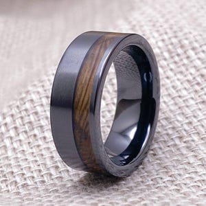 Puede incluir: Un anillo de carburo de tungsteno negro con una incrustaci&oacute;n de madera marr&oacute;n. El anillo tiene un acabado cepillado y un borde pulido.