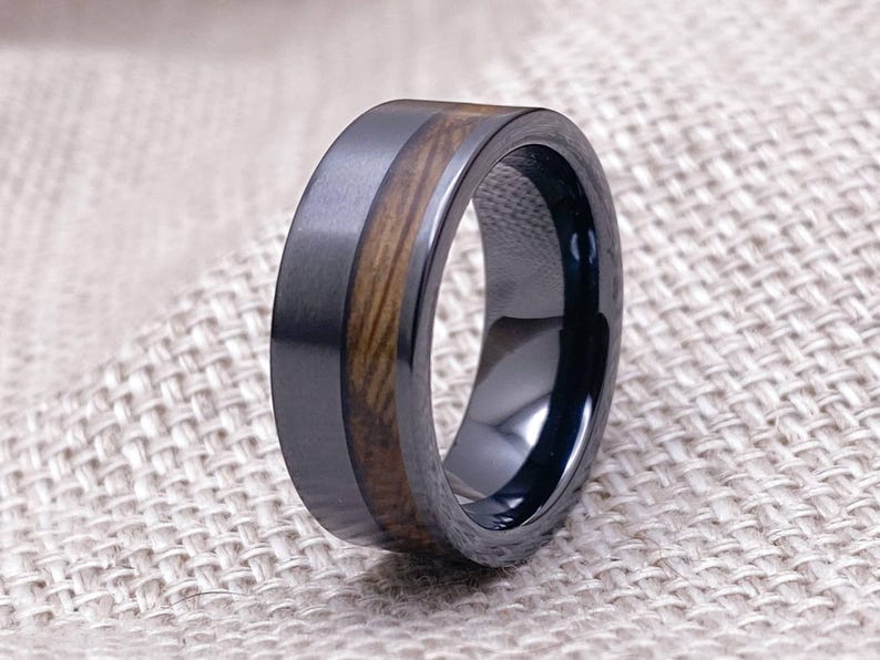 Puede incluir: Un anillo de carburo de tungsteno negro con una incrustaci&oacute;n de madera marr&oacute;n. El anillo tiene un acabado cepillado y un borde pulido.