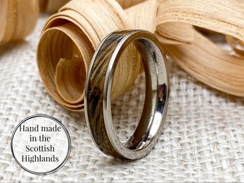 Peut inclure: Gros plan d'une bague argent&eacute;e avec incrustation de bois. La bague est mise en valeur sur un fond de copeaux de bois et une surface textur&eacute;e. Le texte "Hand made in the Scottish Highlands" est dans un cercle.
