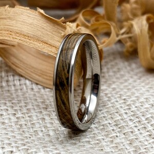 Peut inclure: Gros plan sur une bague avec incrustation de bois et un anneau argent&eacute;. Le bois pr&eacute;sente un motif de grain naturel avec des nuances de marron et de noir. La bague est pos&eacute;e sur un fond neutre avec des copeaux de bois.