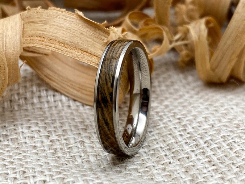 Peut inclure: Gros plan sur une bague avec incrustation de bois et un anneau argent&eacute;. Le bois pr&eacute;sente un motif de grain naturel avec des nuances de marron et de noir. La bague est pos&eacute;e sur un fond neutre avec des copeaux de bois.
