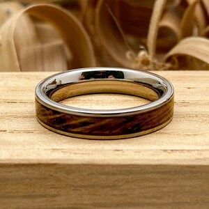 Peut inclure: Une bague en argent et bois avec une finition polie. La bague pr&eacute;sente un anneau en argent avec une incrustation en bois. Le bois a un motif de grain brun fonc&eacute;. La bague est pos&eacute;e sur un bloc de bois.