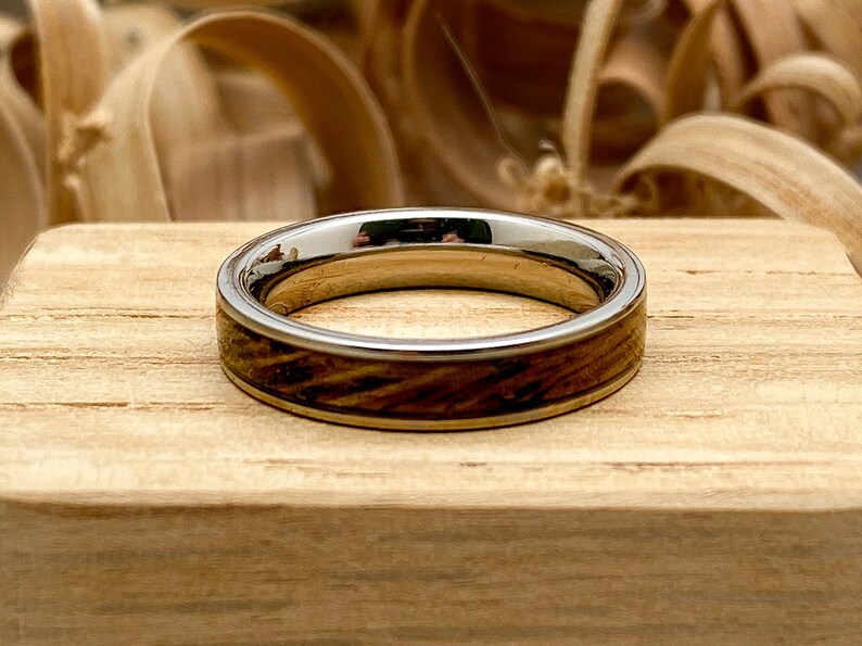 Peut inclure: Une bague en argent et bois avec une finition polie. La bague pr&eacute;sente un anneau en argent avec une incrustation en bois. Le bois a un motif de grain brun fonc&eacute;. La bague est pos&eacute;e sur un bloc de bois.