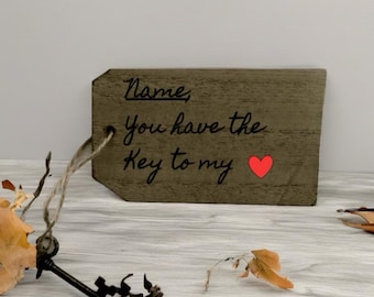 May include: A wooden tag with the words "Name, You have the Key to my" in black script, and a red heart. The tag has a string for hanging. Decorative leaves and a key are in the foreground.