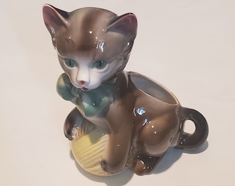 May include: A ceramic planter shaped like a brown and white cat holding a ball of yarn. The cat has green eyes and a pink nose. The planter has a hole in the top for planting.