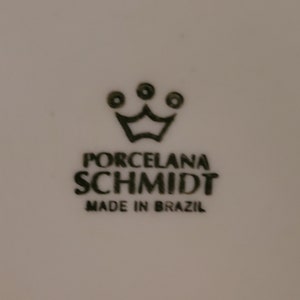 May include: Black and white logo for Porcelana Schmidt, a brand of porcelain. The logo features a crown with three dots above it. The text "Porcelana Schmidt" is written in all caps, with "Made in Brazil" below it.