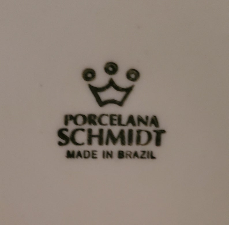 May include: Black and white logo for Porcelana Schmidt, a brand of porcelain. The logo features a crown with three dots above it. The text "Porcelana Schmidt" is written in all caps, with "Made in Brazil" below it.