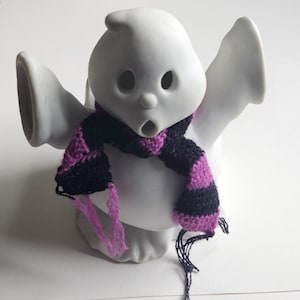 May include: A white ceramic ghost figurine with hollow eyes and an open mouth, wearing a small, hand-knitted scarf featuring horizontal stripes in alternating black and purple yarn with loose fringe ends hanging down the front of the figure.