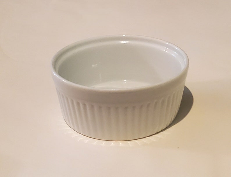May include: A white ceramic ramekin with a fluted design. The ramekin is empty and ready to be used for baking or serving.