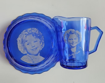 May include: A blue glass pitcher and plate set. The pitcher has a handle and a portrait of a smiling child. The plate is round with a scalloped edge and also features the child's portrait. The set is a vibrant blue.
