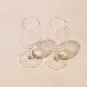 Puede incluir: Dos copas de vino de vidrio transparente con tallo y base redonda. Las copas están vacías y están sobre una superficie blanca.