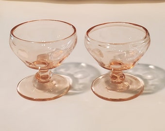 May include: Two pink glass dessert dishes with a scalloped rim and a textured base. The dishes are on a white surface.