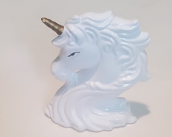 May include: A white ceramic unicorn head figurine with a gold horn. The unicorn has a flowing mane and a serene expression. The figurine is a decorative item.