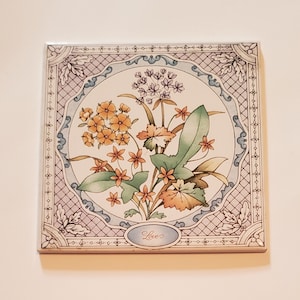 May include: A square ceramic tile with a floral design. The tile features a bouquet of yellow, orange, and purple flowers with green leaves. The tile has a light blue background with a white border and the word "Love" in the center.