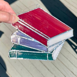 May include: A stained glass ornament in the shape of a stack of books. The top book is red, the middle book is purple, and the bottom book is green. The pages are white, and the books are outlined in silver.