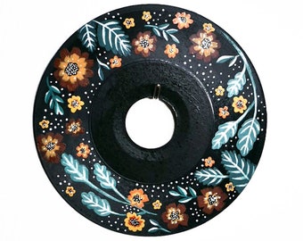 May include: A black vinyl record painted with a floral design in shades of orange, brown, and teal. The record is hanging on a white wall. A vintage camera and a framed piece of wood with the text "ACTUAL PIECE OF STAGE FROM THE WOODSTOCK FESTIVAL AUGUST 15-18, 1969" are on a wooden surface.
