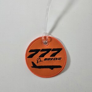 May include: Round orange luggage tag with the number "777" and the word "BOEING" in black. The tag has a clear plastic loop for attachment.