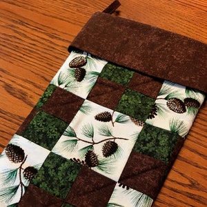 Könnte beinhalten: Ein brauner und grüner Weihnachtsstrumpf mit einem karierten Muster und Tannenzapfendesigns. Der Strumpf hat einen braunen Filz-Manschetten.