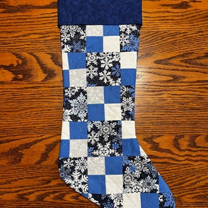 Puede incluir: Una media de Navidad acolchada azul y blanca con un patr&oacute;n de copo de nieve. La media tiene un pu&ntilde;o azul y un lazo para colgar.