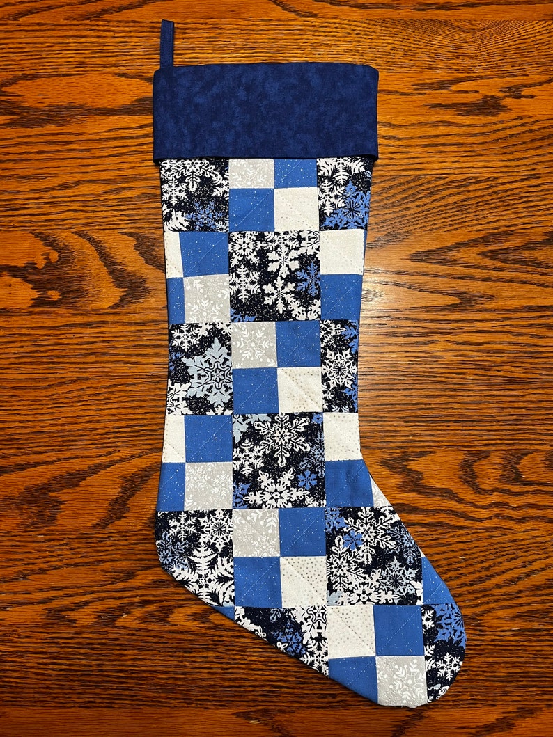 Puede incluir: Una media de Navidad acolchada azul y blanca con un patr&oacute;n de copo de nieve. La media tiene un pu&ntilde;o azul y un lazo para colgar.