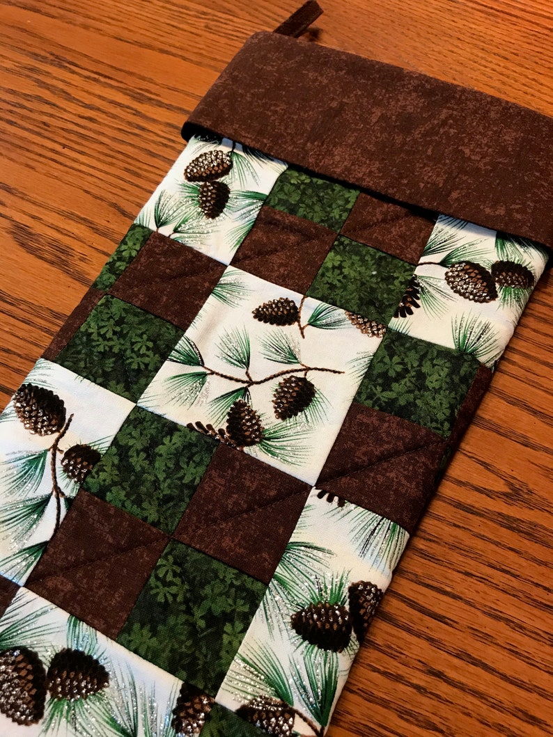 May include: A brown and green Christmas stocking with a checkered pattern and pine cone designs. The stocking has a brown felt cuff.