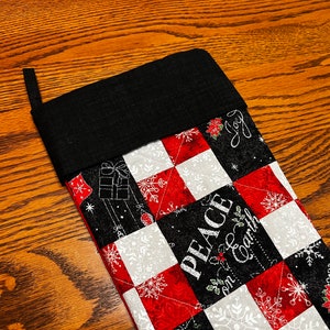 Könnte beinhalten: Ein schwarz-weißer Weihnachtssocken mit rotem und weißem Patchwork-Muster. Der Socken trägt den Text "Peace on Earth" und "Joy".