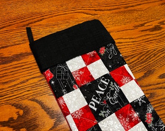 Pode incluir: Uma meia de Natal preta e branca com um design de patchwork vermelho e branco. A meia tem o texto "Peace on Earth" e "Joy".