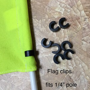 May include: Black plastic flag clips for a 1/4 inch pole. The clips are designed to hold a flag securely to a pole. The text "Flag clips fits 1/4" pole" is written below the clips.