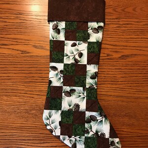 May include: A brown and green Christmas stocking with a pine cone and branch pattern. The stocking has a brown cuff and a white background.
