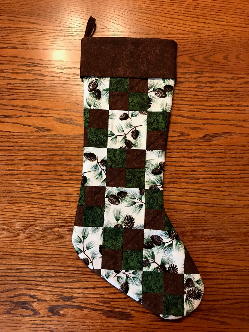 May include: A brown and green Christmas stocking with a pine cone and branch pattern. The stocking has a brown cuff and a white background.