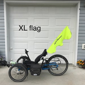May include: A black recumbent trike with a yellow flag that says "Full Fat Adventurer" attached to the back. The trike is parked in front of a garage door with the words "XL flag" written above the door.