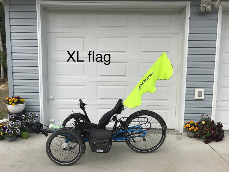 May include: A black recumbent trike with a yellow flag that says "Full Fat Adventurer" attached to the back. The trike is parked in front of a garage door with the words "XL flag" written above the door.