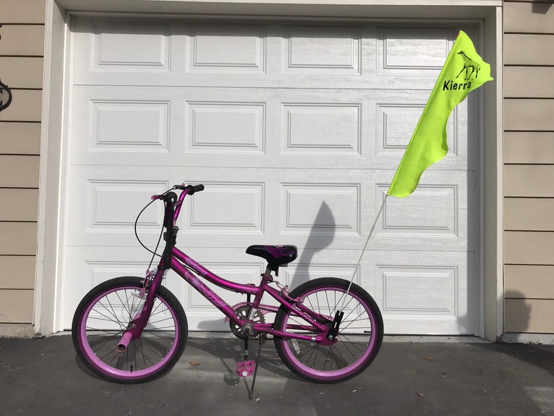 May include: A purple bicycle with pink wheels and a yellow flag with the name "Kierra" on it.