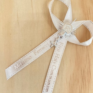 May include: White satin ribbon with a silver charm featuring a chalice and the letters "IHS". The ribbon is tied in a bow and has the text "First Holy Communion" and "Massimo Pettinelli" printed on it.