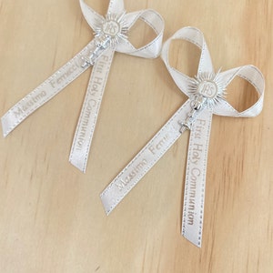 May include: Two white satin ribbon bows with silver charms and the text "First Holy Communion" and "Massimo Ferrucci" printed on the ribbons.