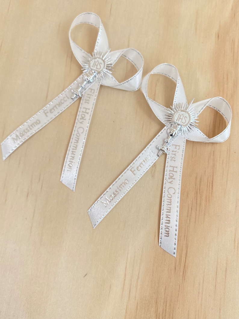 May include: Two white satin ribbon bows with silver charms and the text "First Holy Communion" and "Massimo Ferrucci" printed on the ribbons.