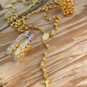 May include: A gold rosary with a small gold cross and a gold medal charm. The rosary is displayed on a wooden surface with two small glass bottles filled with gold beads.