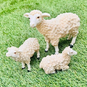 May include: Three white fluffy sheep figurines, one standing and two sitting, on a green artificial grass background.